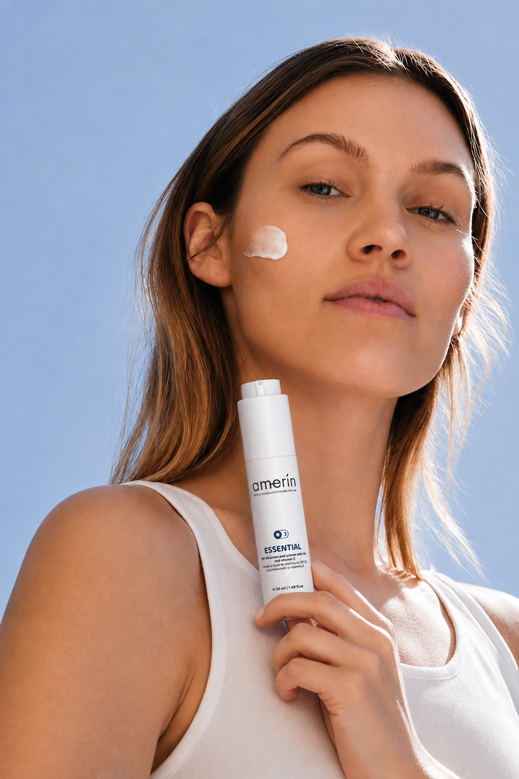 A woman with a dollop of white cream on her cheek holds a white ESSENTIAL | SPF30 Cream and Primer with HA and Vitamin E skincare bottle against a clear blue sky background.