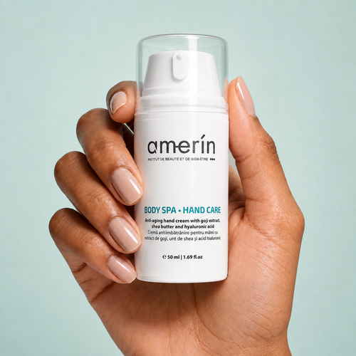 A hand with manicured nails holds a white Amerín anti-aging hand cream pump bottle against a light blue background.